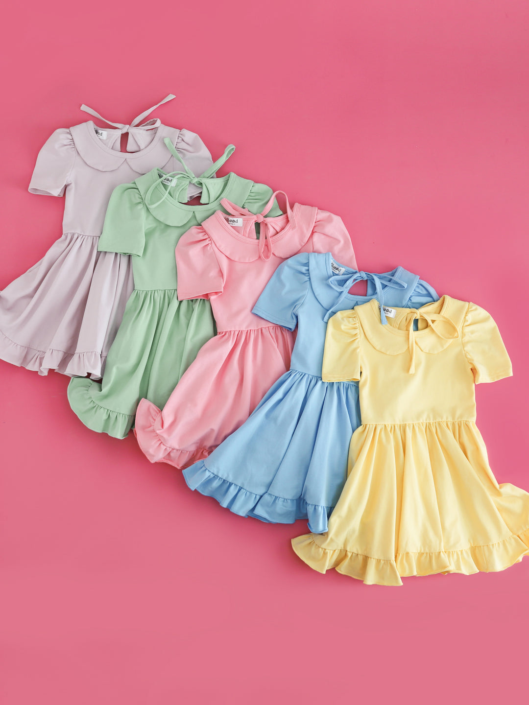 flat lay of five solid, cotton blend, pastel twirl dresses with peter pan collars for girls' spring collection in lavender, mint, blossom pink, bluebell and sunshine yellow