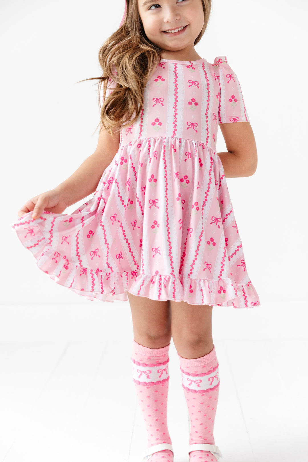 girl wearing short sleeve twirl dress in pink with wallpaper print featuring bows and flowers for Easter with matching pink knee highs with dots and printed bows