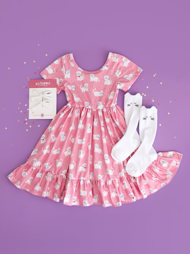 flatlay of girls pink short sleeve twirl dress with white kitten print with matching white cat knee high socks and white yarn pigtail bows