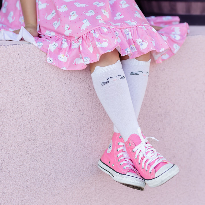 girls legs in white knee highs with cat ears and face and wearing a pink short sleeve twirl dress with white kitten print and pink converse