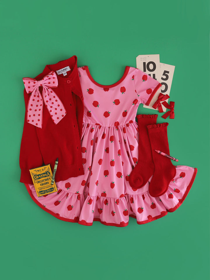 girl's pink apple print twirl dress with red trim paired with matching apple print bow, bright red cardigan and pointelle knee socks