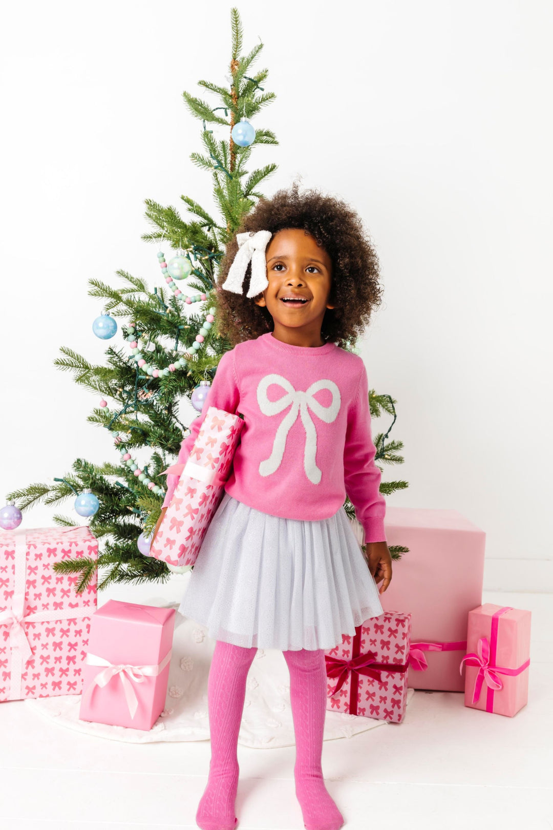 young girl holding Christmas gift in pink sweater with sparkly white bow design paired with matching sparkly white tutu skirt and haor bow and pink cable knit tights