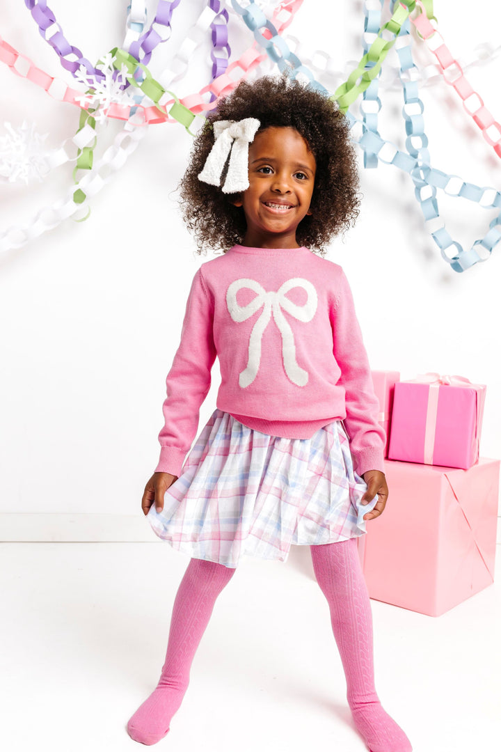 little girl in front of winter wonderland decor with pink bow sweater paired with sparkly white bow, pastel plaid velvet skirt and matching pink cable knit tights