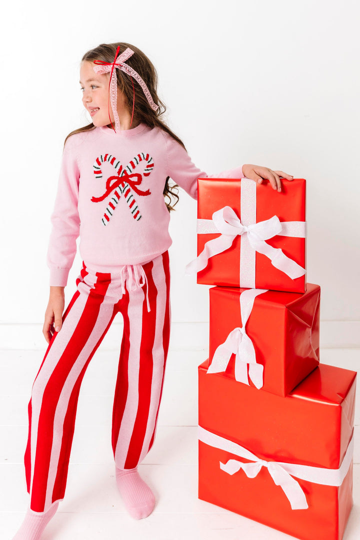 girl by stack of Christmas gifts wearinfgpink pullover sweater with candy cane design and matching red and pink striped sweater pants 