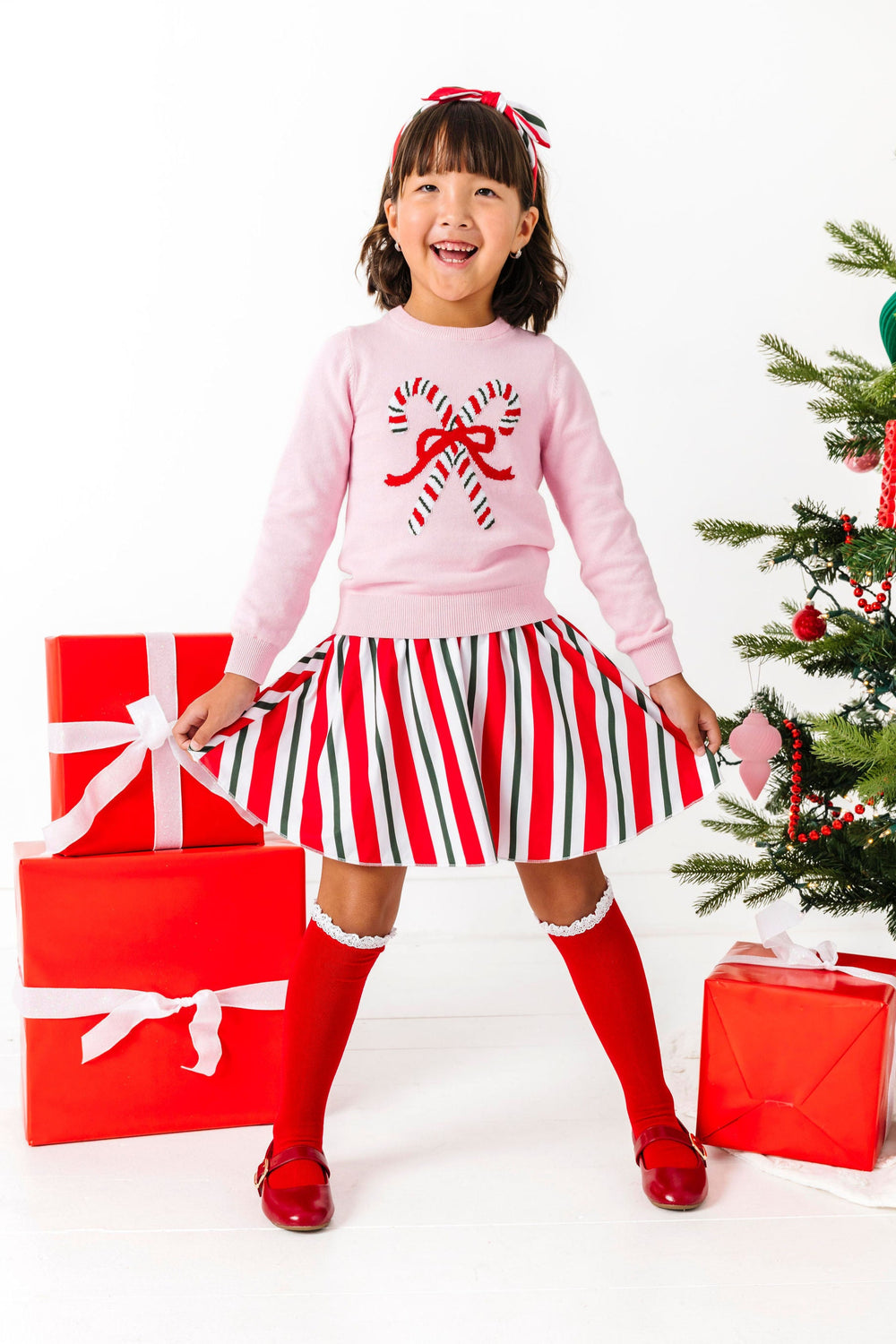 young girl in pink Christmas sweater with red, white and green candy cane design, matching skirt and headband and red knee highs with white lace tirm
