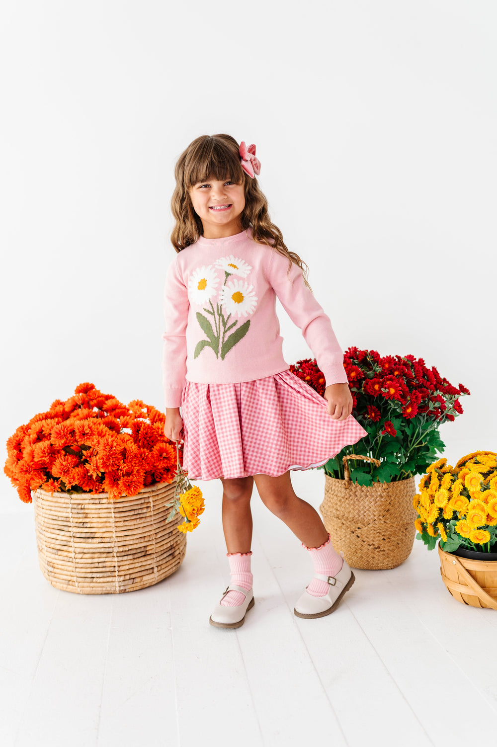 little girl in front of fall flower baskets in pink daisy sweater, pink gingham lettuce trim twirl dress, pink ribbed midi socks and matching velvet hair bow