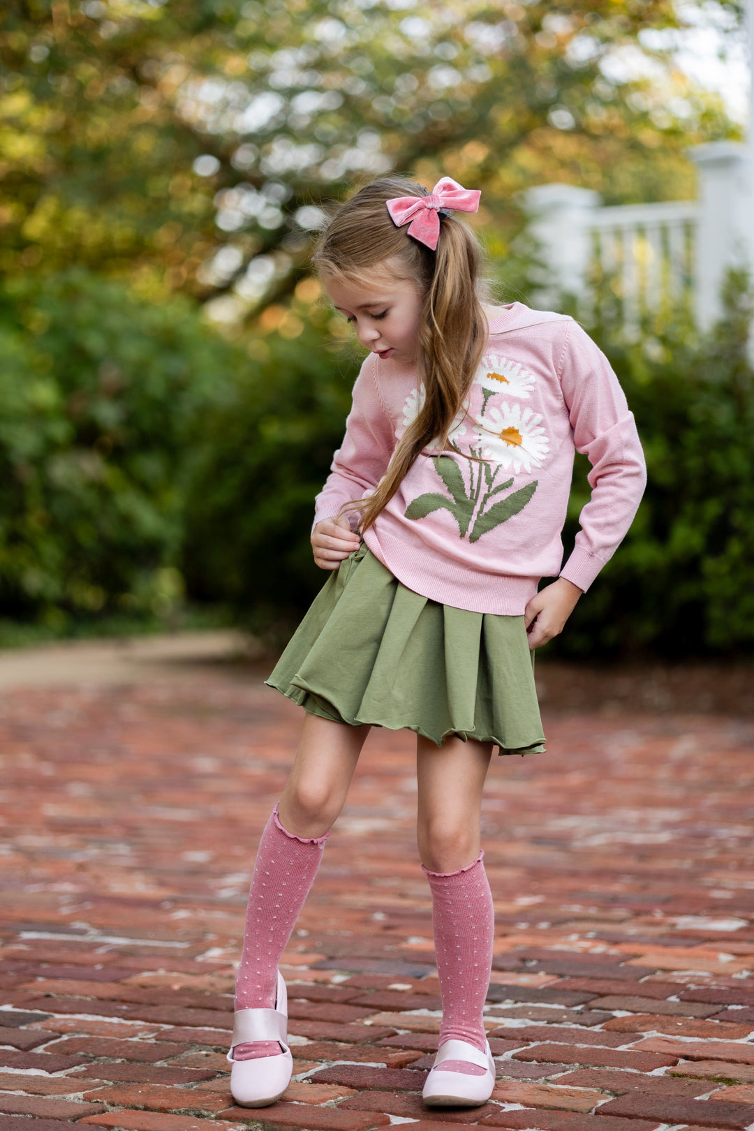 young girl in garden wearing light pink cotton knit sweater with ivory daisy design paired with olive lettuce trim twirl dress, pink velvet bow and matching pink dotted knee socks