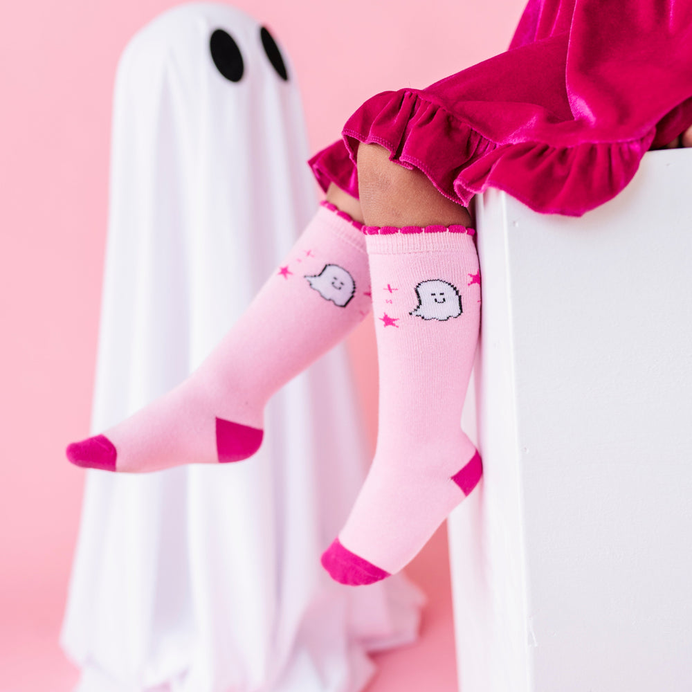 girl sitting on white box with sheet ghost in the background wearing hot pink velvet twirl dress paired with two-tone pink knee socks with ghost design 