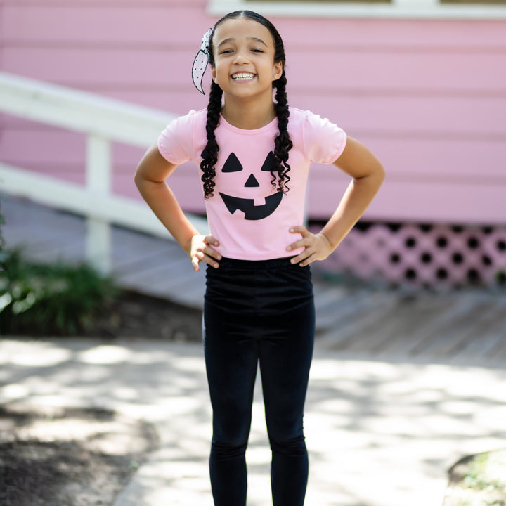 little girl in pink fitted top with black jack-o-lantern face and black velvet leggings for Halloween