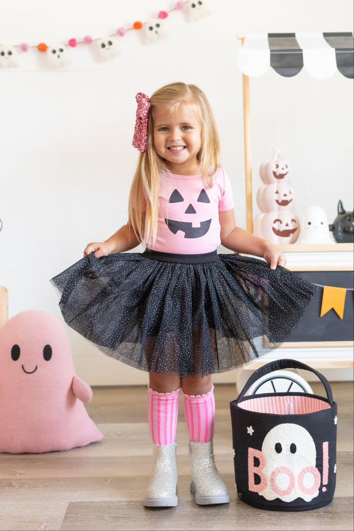 little girl in front of Halloween decor with pink pumpkin tee black sparkly tulle skort, pink striped knee highs and pink sequin bow for pinkoween