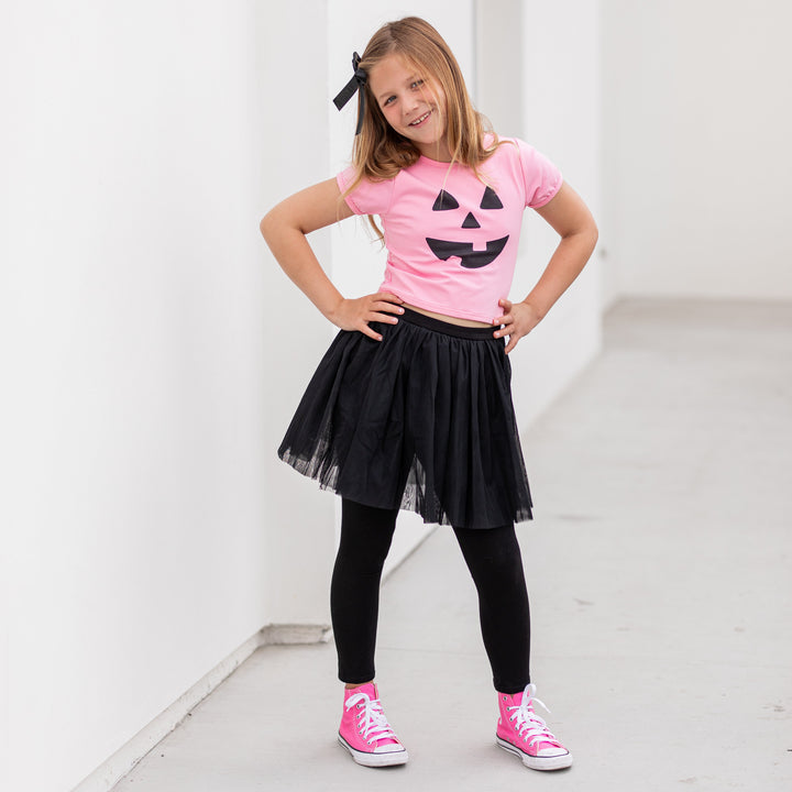 girl in pink tee with black jack-o-lantern face, black leggings with attached tulle skirt, black shimmery ribbon bow and pink high tops