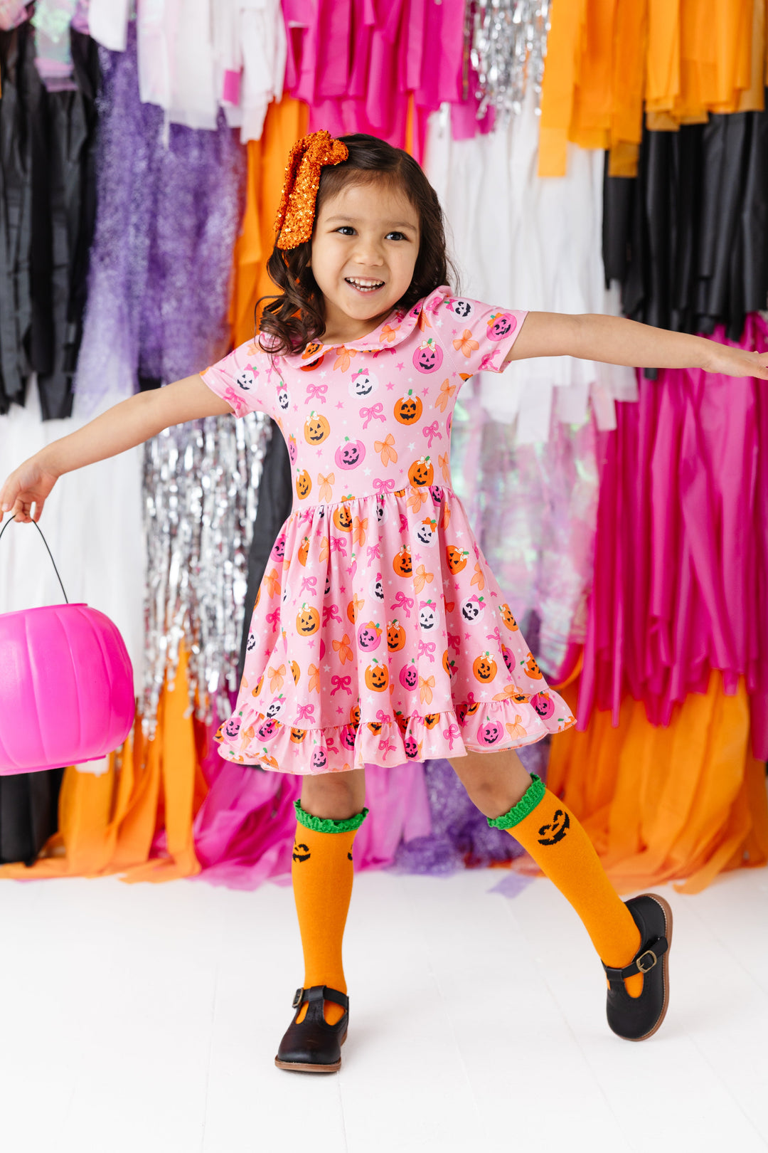 little girl holding pink pumpkin bucket in pink short sleeve collared twirl dress with pumpkins wearing bows and matching orange sequin bow and pumpkin knee socks