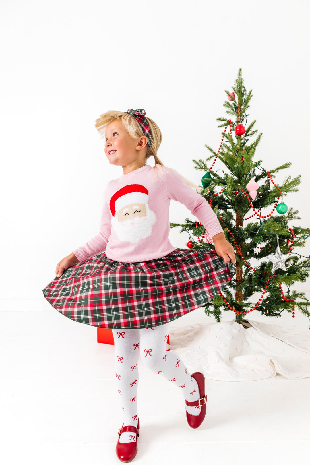 little girl twirling by Christmas tree in pink knit Santa sweater with fluffy beard, Christmas plaid skirt and headband and white knit tights with red bow pattern