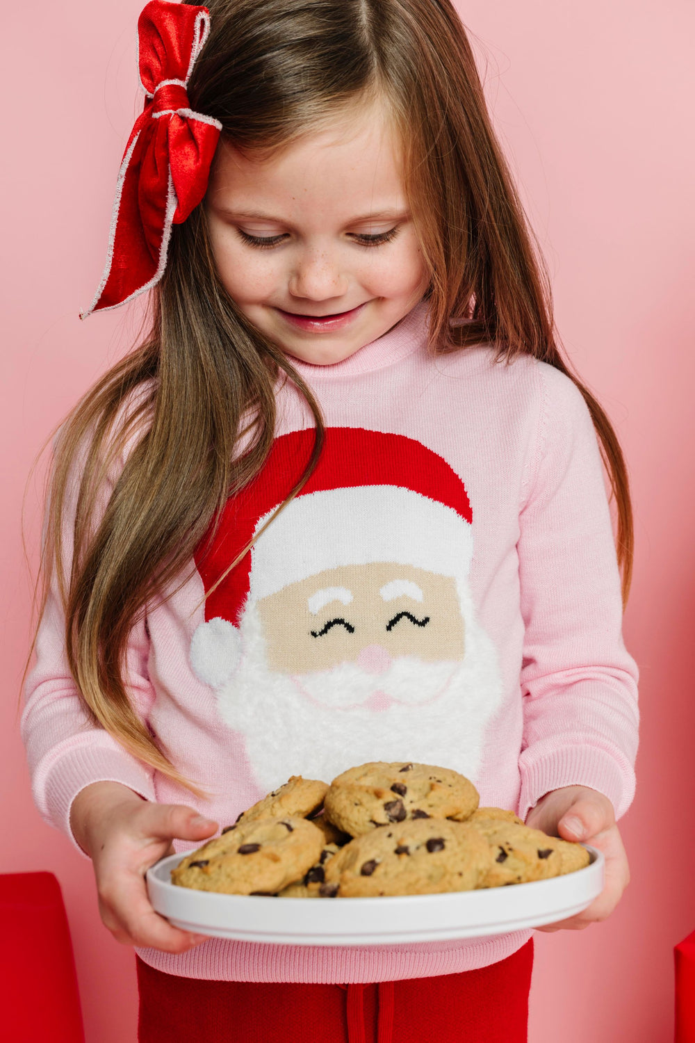 little girl holding tray of cookies in pink Santa sweater with fluffy white beard and matching red velvet bow with white trim