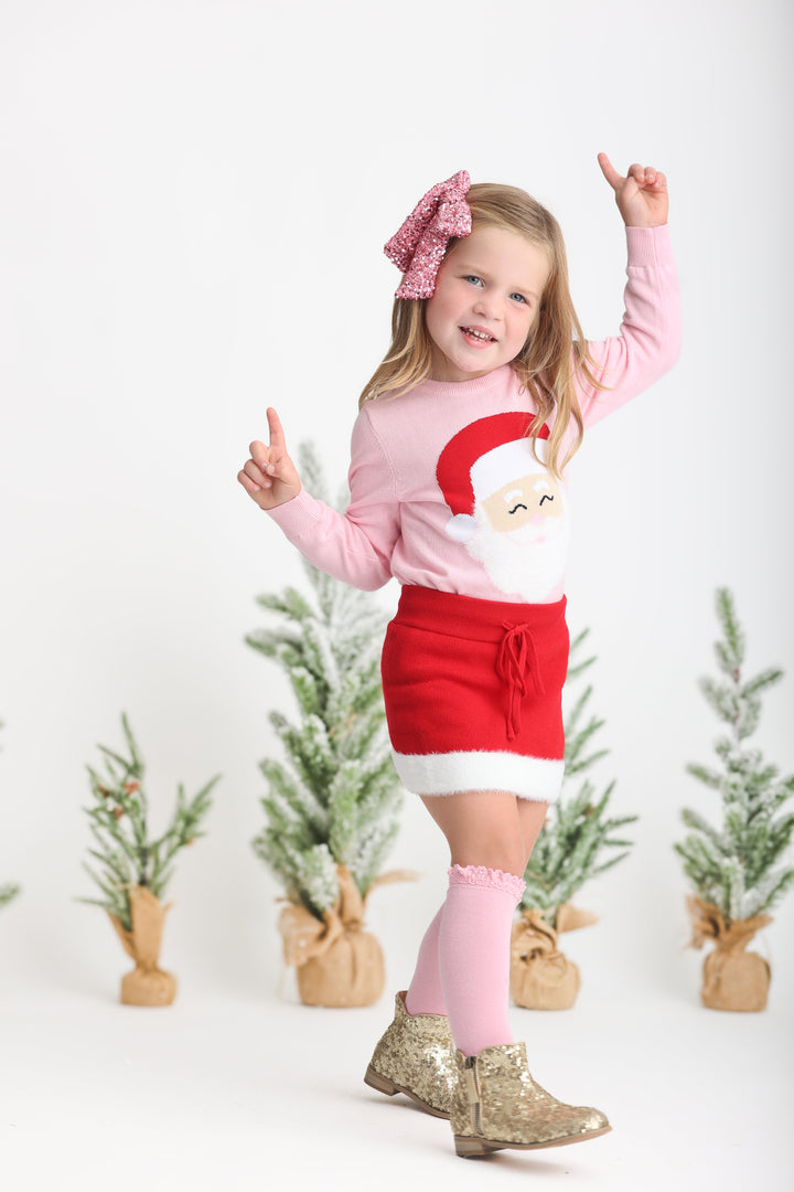 little girl dancing in front of Christmas trees in pink Santa sweater with matching sweater skirt and sparkly pink bow