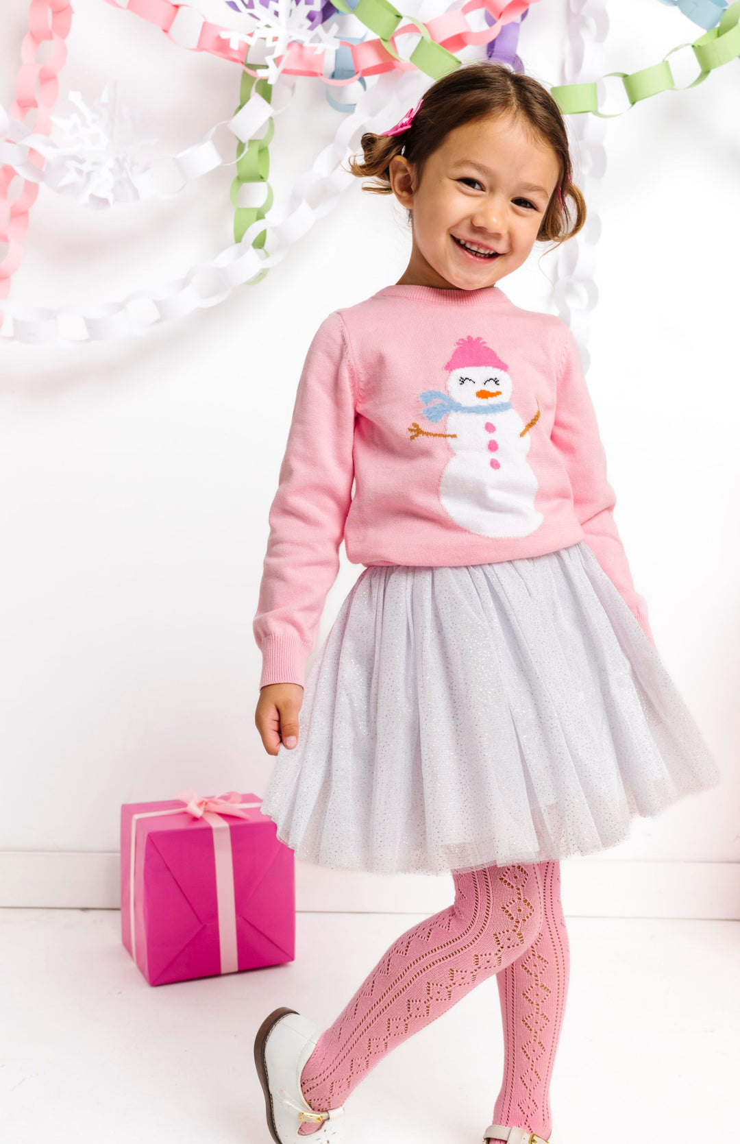 little girl in winter wonderland wearing pink pullover sweater with snowgirl design, white sparkly tutu skirt and matching open knit pink tights