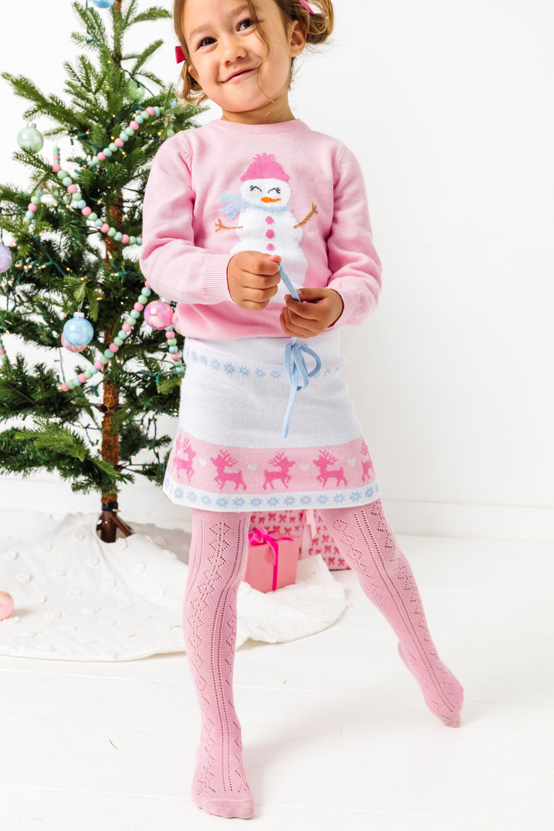 young girl in front of Christmas tree in pink pullover sweater with snowman design paired with white fair isle print sweater skirt and pastel pink open knit tights