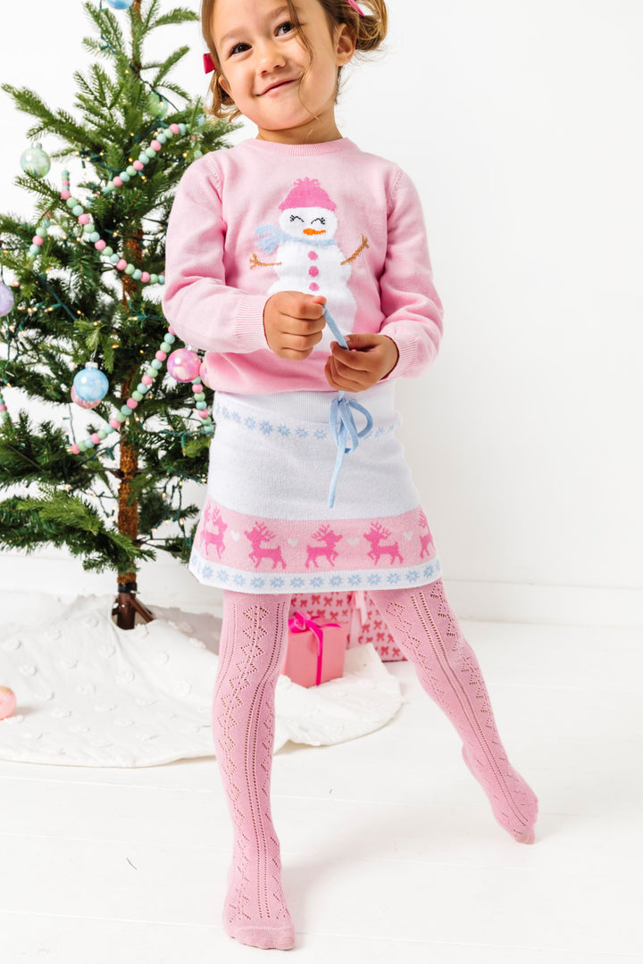 young girl in front of Christmas tree in pink pullover sweater with snowman design paired with white fair isle print sweater skirt and pastel pink open knit tights