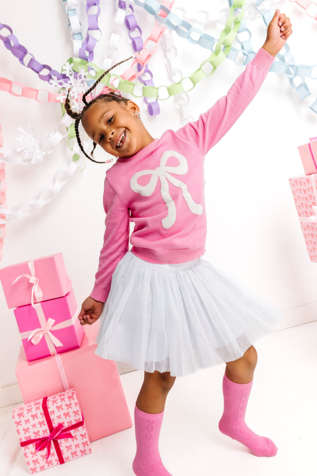 little girl dancing in front of pink Christmas gifts in pink knit sweater with sparkly white bow design, matching sparkly white tutu and pink cable knit knee highs