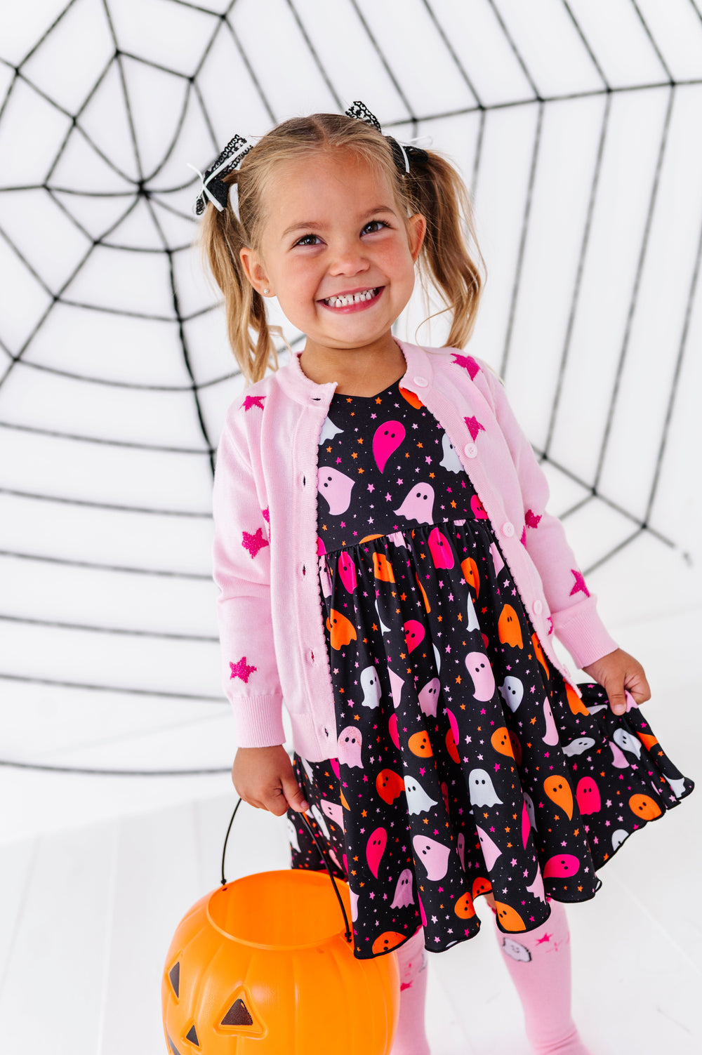 young girl holding pumpkin bucket in vibrant ghost print twirl dress paired with pink sparkle star cardigan and matching pink ghost knee socks