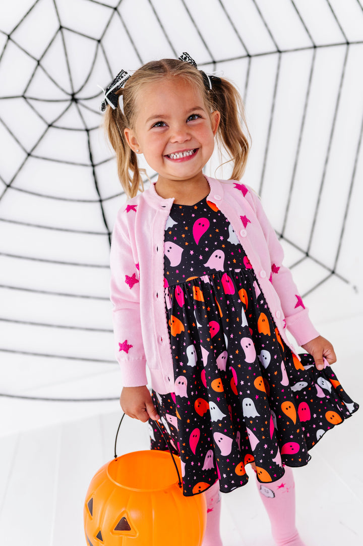 young girl holding pumpkin bucket in vibrant ghost print twirl dress paired with pink sparkle star cardigan and matching pink ghost knee socks