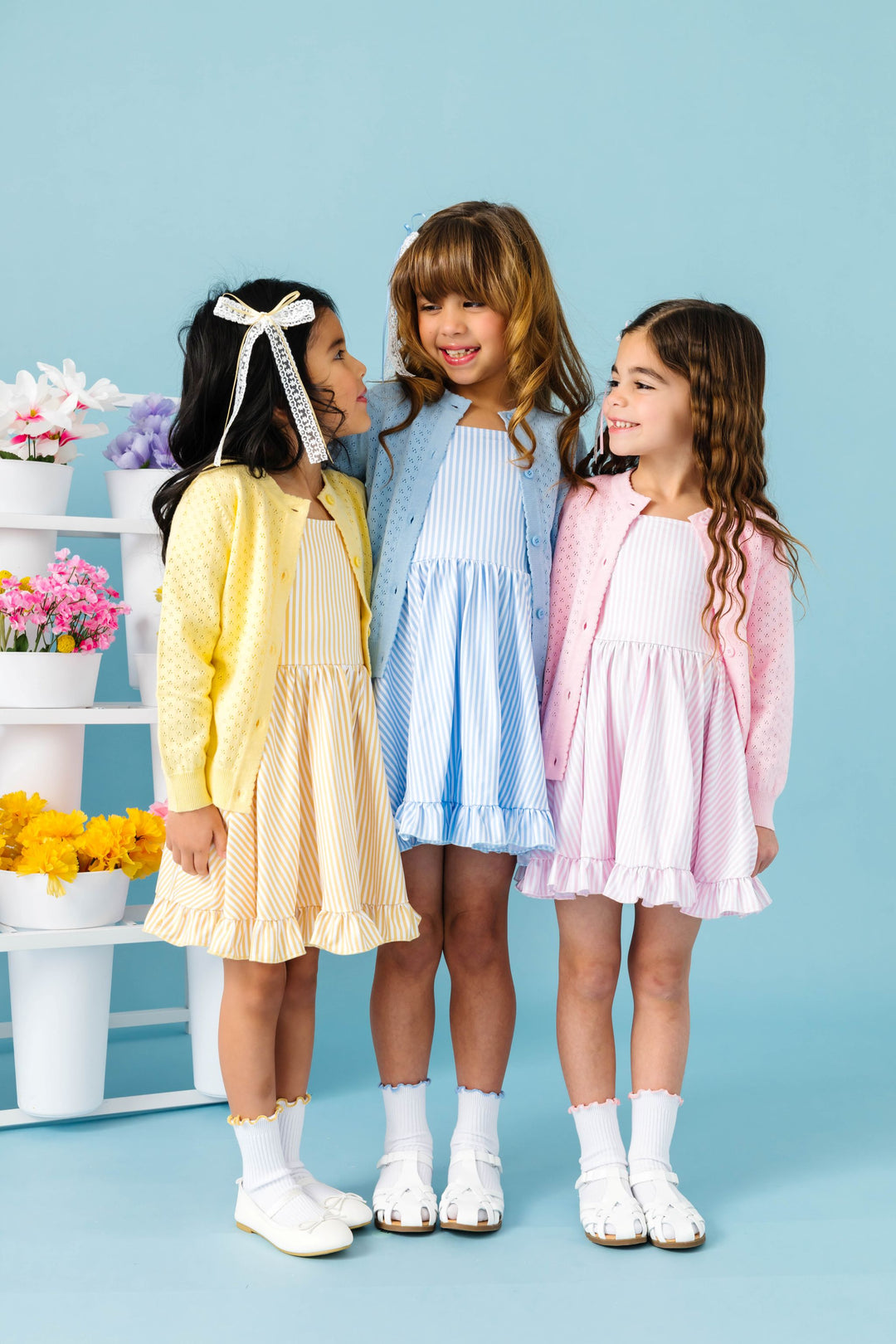 three girls wearing pinstripe print spring twirl dresses in pastel yellow, light blue and pink with matching pointelle cardigans, hair bows and midi socks with lettuce trim
