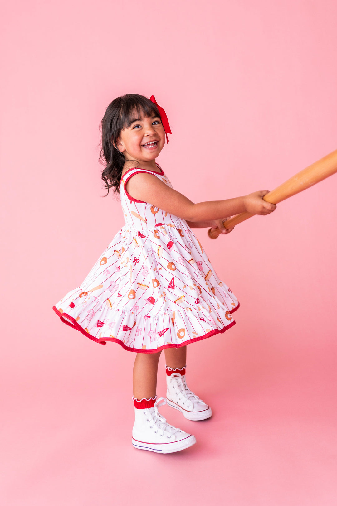little girl twirling with baseball bat in baseball themed tank twirl dress, red satin bow and red midi socks