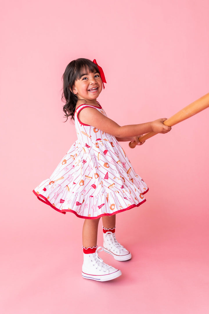 little girl twirling with baseball bat in baseball themed tank twirl dress, red satin bow and red midi socks