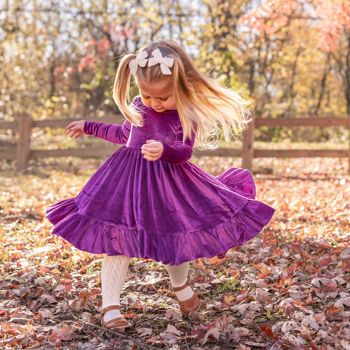 little girl twirling in fall leaves wearing long sleeve purple velvet twirl dress, cream pointelle knit tights and cream velvet pigtail bows