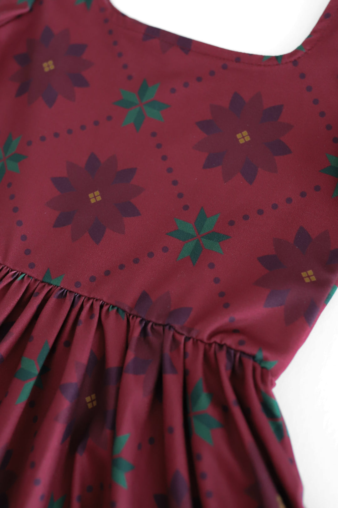 Close-up of a sleeveless dress or top in deep burgundy with a repeating poinsettia floral and starburst print in dark purple and green, plus small dotted lines, against a white background.
