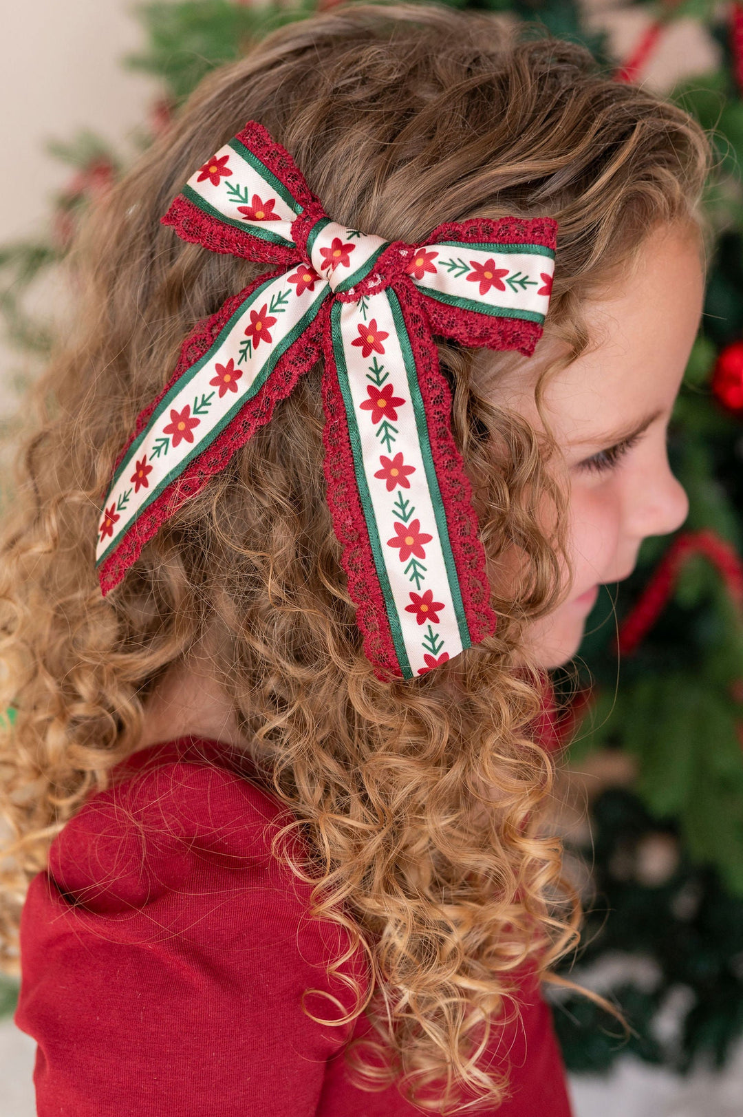 A young girl with curly blonde hair wearing a red dress and a large red, green, and cream poinsettia patterned bow with lace trim, standing in front of a decorated Christmas tree.