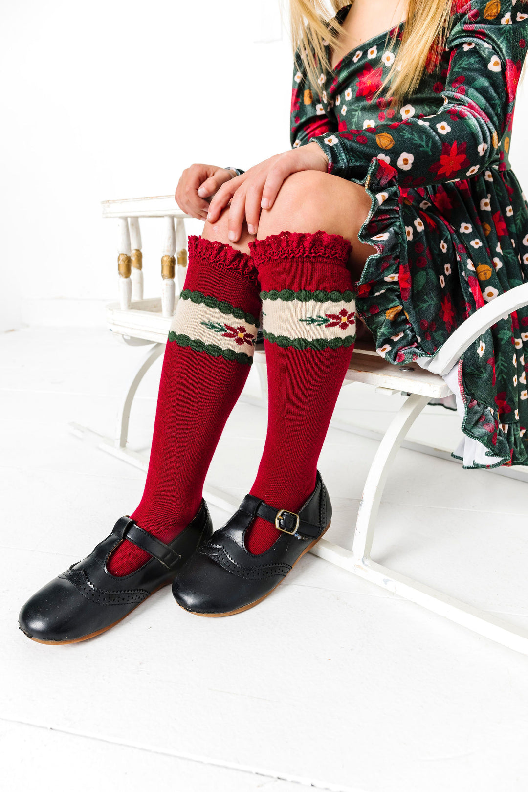 girl sitting on sled in crimson knee highs with poinsettia design, black mary janes and matching velvet poinsettia print dress 