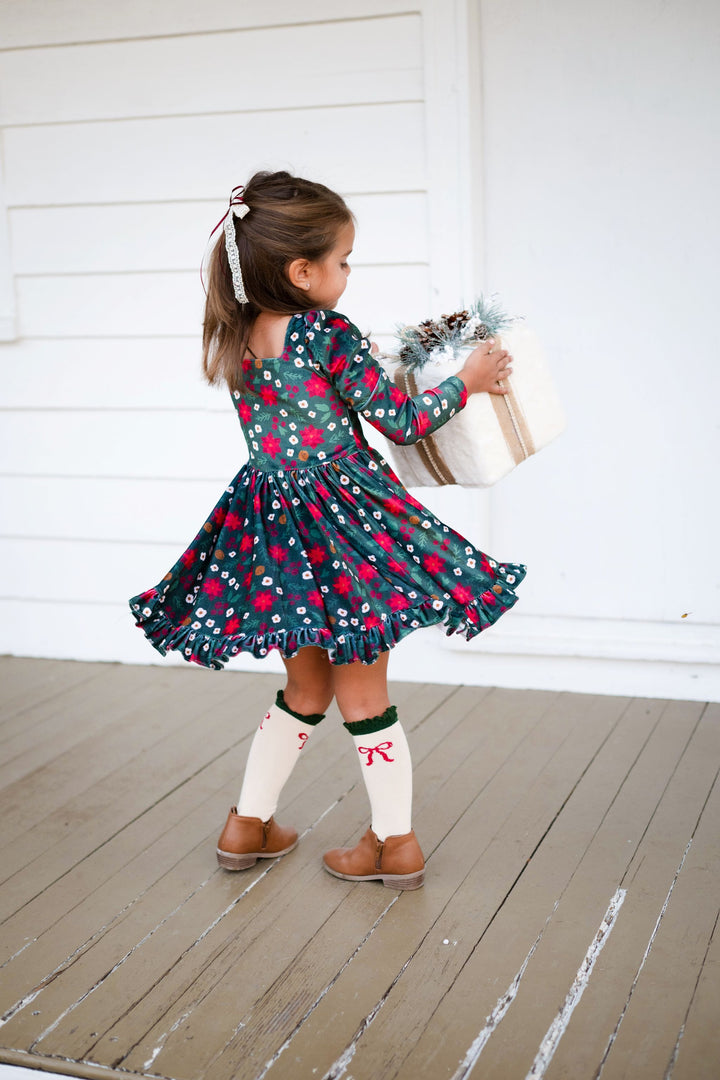little girl twirling with Christmas gift in forest green velvet dress with poinsettia print paired with cream knee highs with crimson bow design and forest green lace top