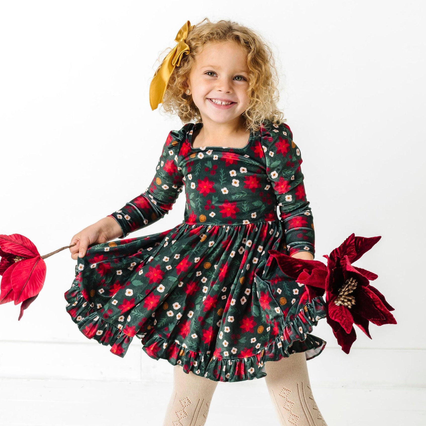 little girl holding poinsettias in green velvet long sleeve twirl dress with poinsettia print, gold satin bow and open knit cream tights for Christmas party