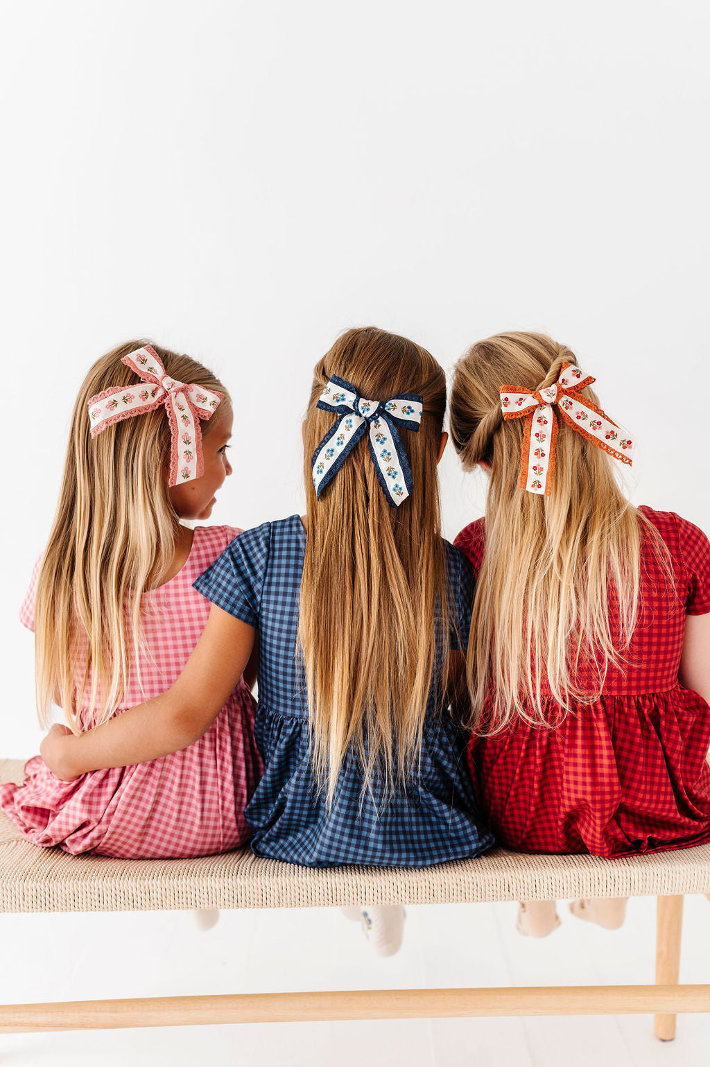 Three young girls sitting side by side on a woven bench, viewed from behind, wearing matching fall gingham dresses and decorative hair bows in pink, blue, and rust with floral patterns.