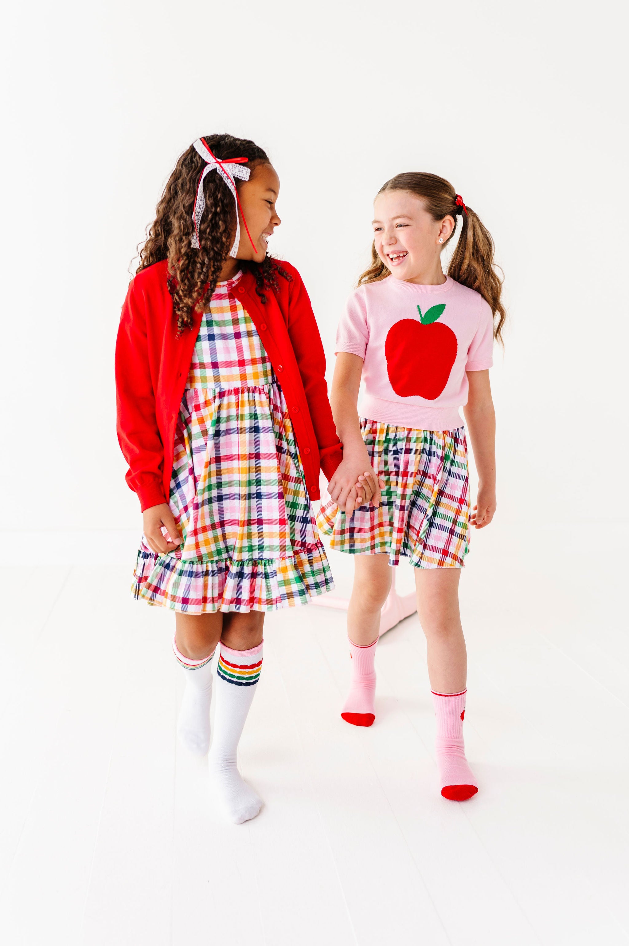 girls holding hands in primary colored gingham dress and skort paired with apple sweater and bright red cardigans