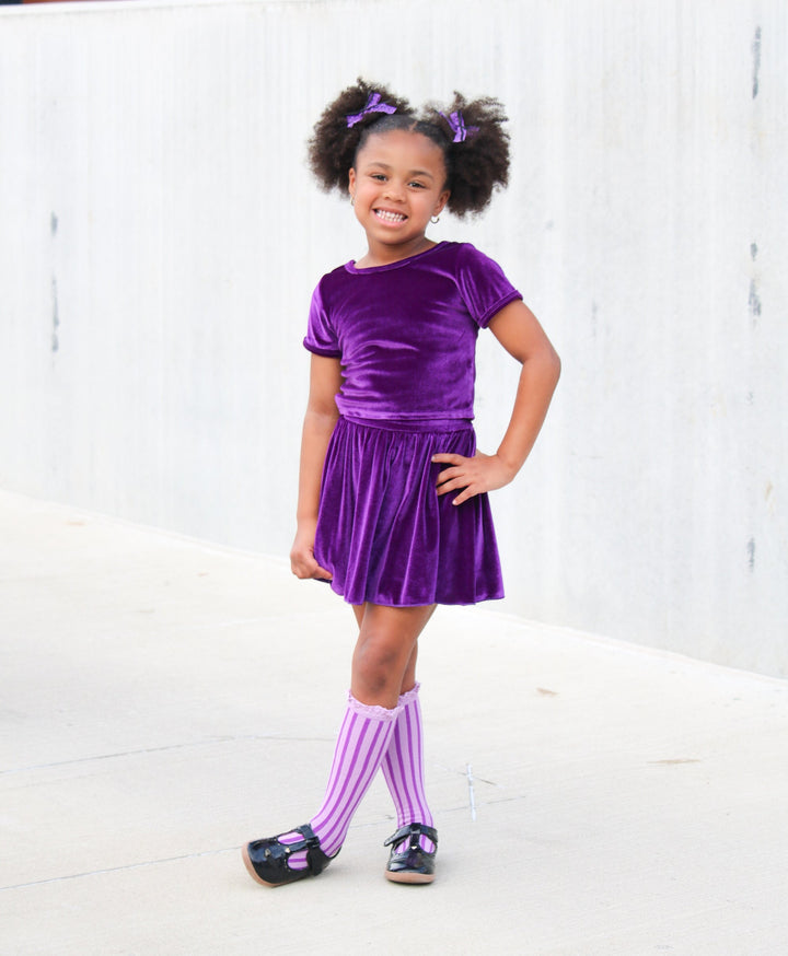 little girl in purple lace pigtails, matching top and skorts in purple velvet and two-tone striped purple knee socks and black mary janes for Halloween vibes