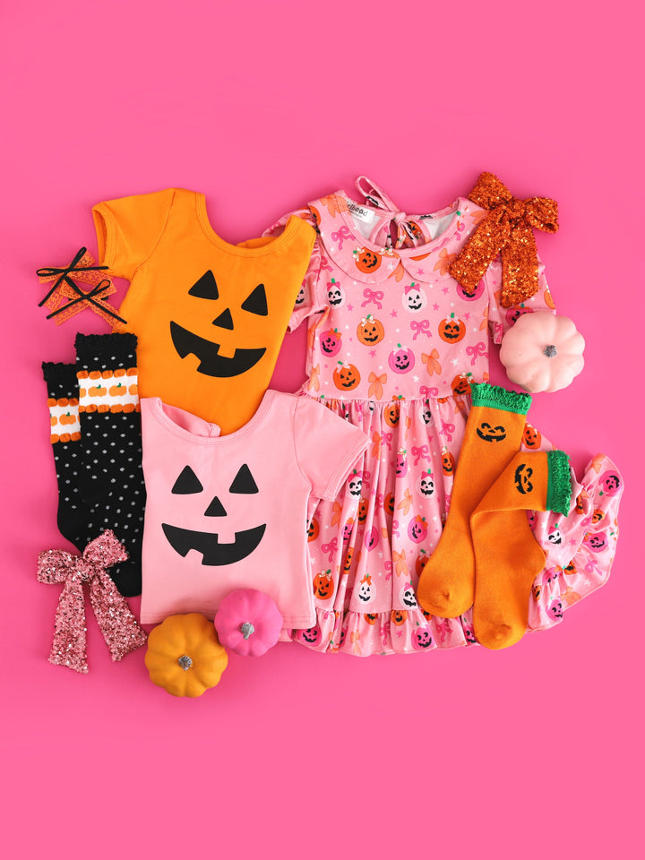 flat lay with Halloween pumpkin face tees in pink and orange, pink pumpkin printed twirl dress and matching socks and bows 