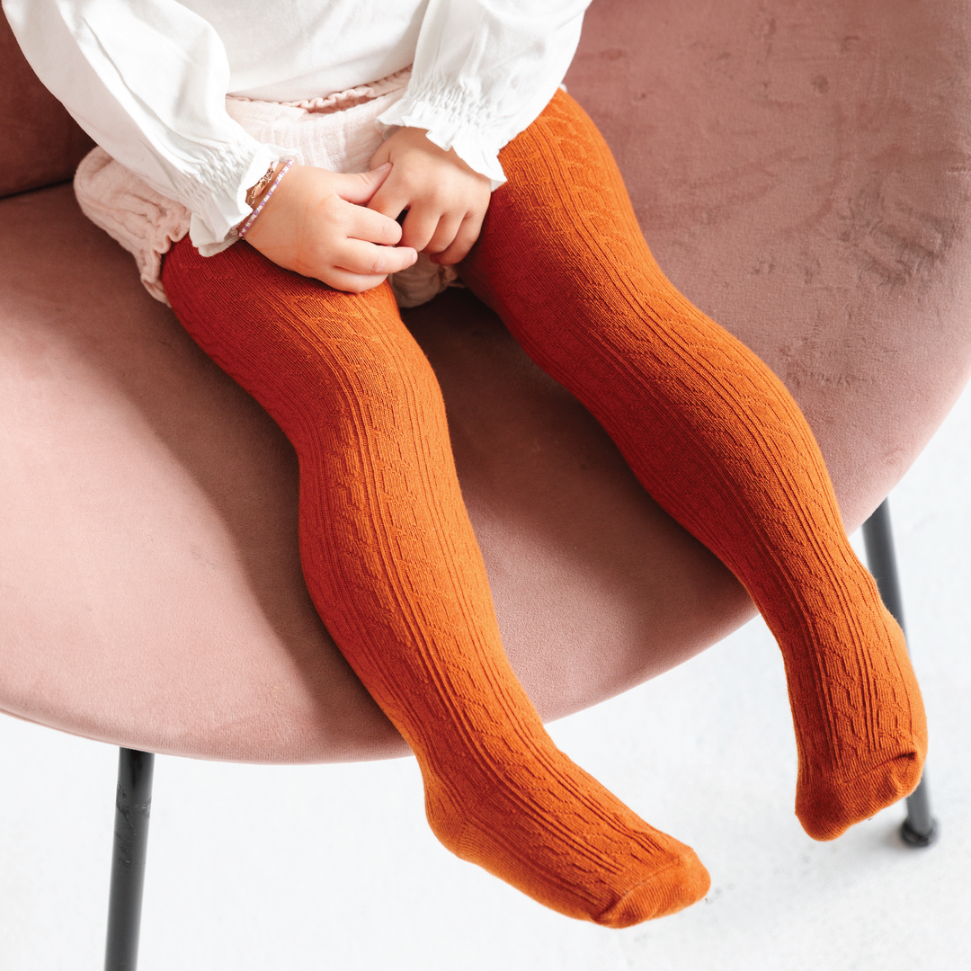 toddler girl wearing pumpkin spice orange cable knit tights on pink chair and white outfit