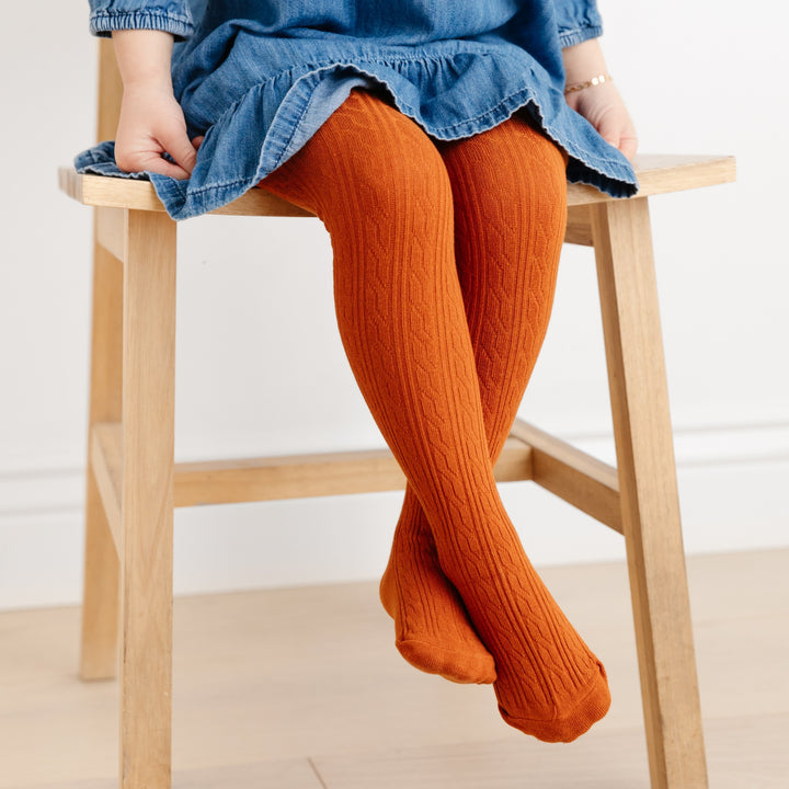 girl in denim dress sitting in wooden chair wearing fall orange pumpkin spice cable knit tights