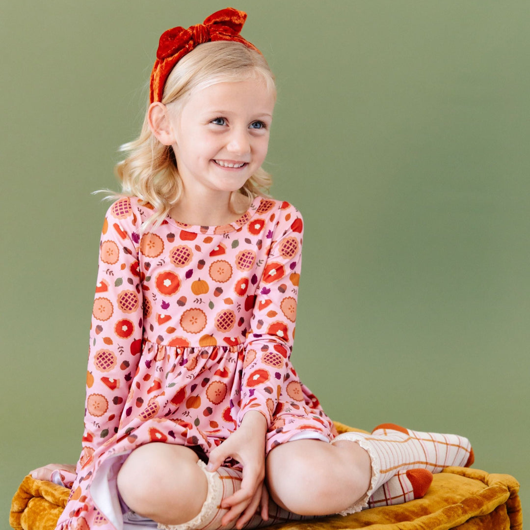 little girl in pumpkin pie printed long sleeve dress with matching orange velvet headband and grid patter knee highs