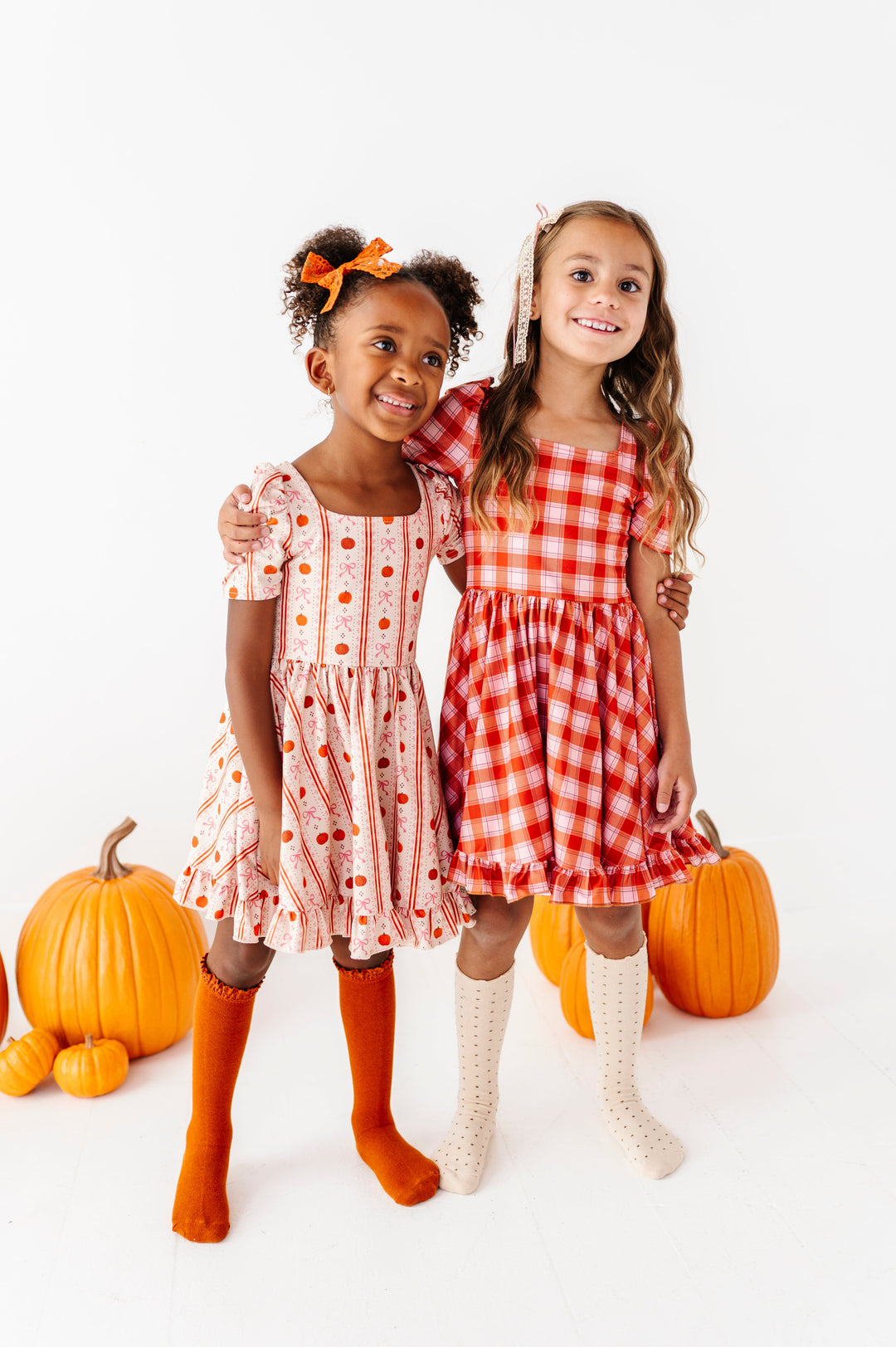 twirl in harvest themed outfits, one wearing cream twirl dress with pumpkin and bow vintage wallpaper inspired print with burnt orange accessories and the other in orange plaid square neck twirl dress paired with cream accessories
