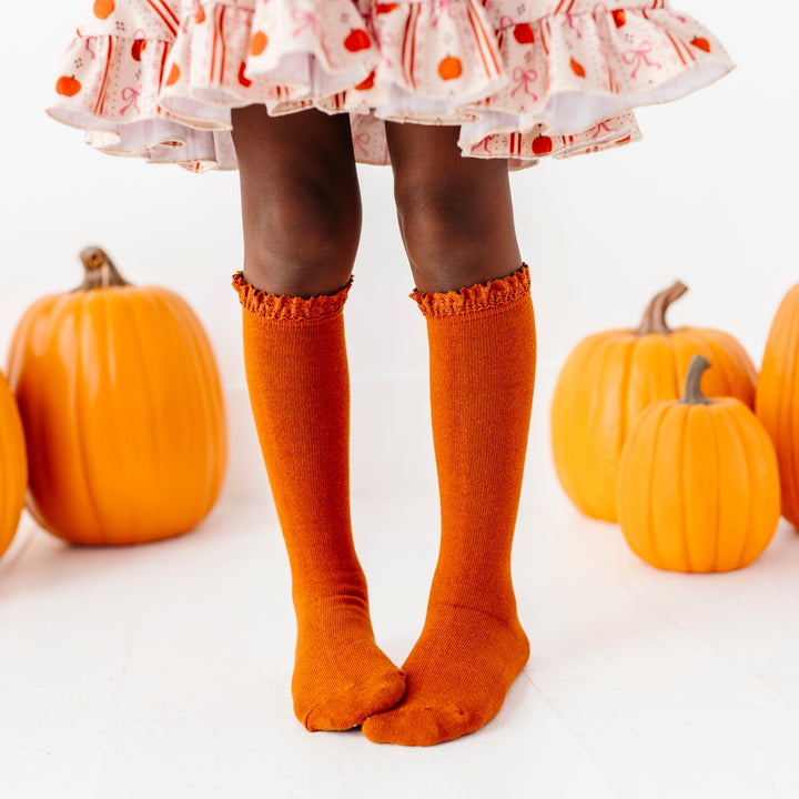 little girl in front of orange pumpkins in pumpkin wallpaper inspired twirl dress paired with matching pumpkin spice lace top knee socks