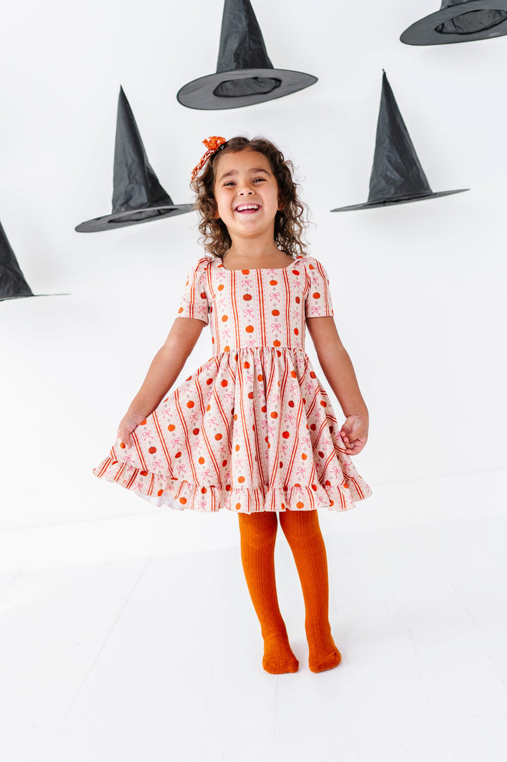 little girl posing in front of witch hats in cream short sleeve twirl dress with vintage pumpkin and bow wallpaper print and orange cable knit tights to match