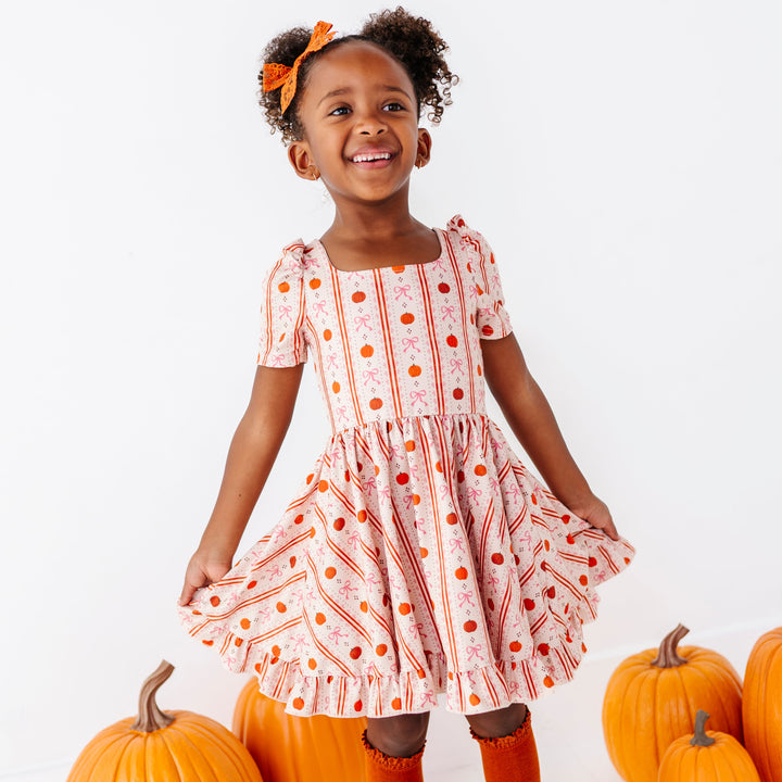 little girl posing in front of pumpkins in cream short sleeve twirl dress with pretty vintage pumpkin and bow wallpaper print paired with matching orange knee socks and lace bow