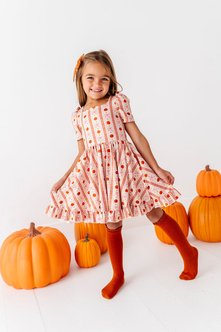 A young girl wearing a short-sleeve dress with a pumpkin and striped pattern, standing in a studio surrounded by pumpkins. She is wearing orange knee-high socks and an orange bow in her hair.