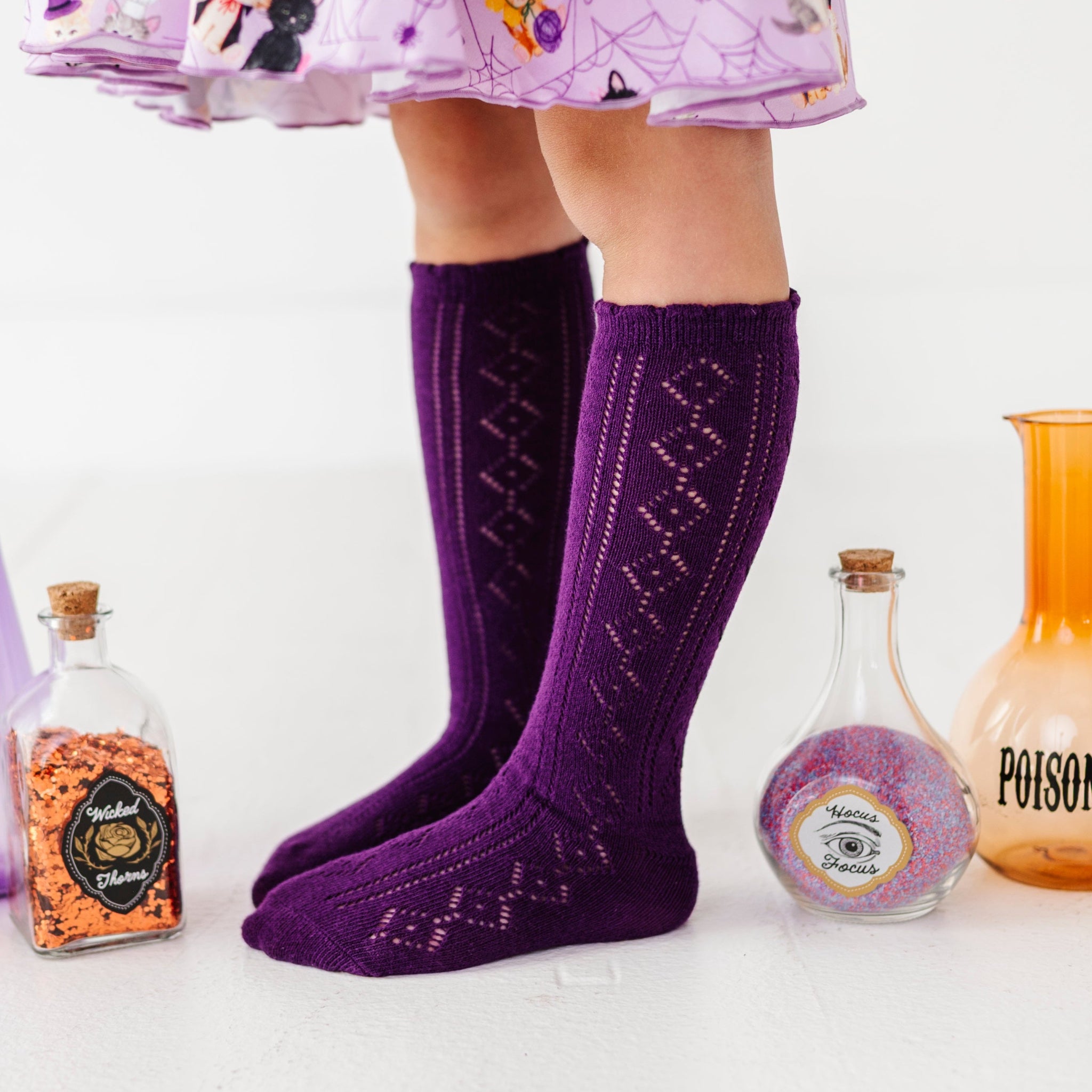 little girl in deep purple open knit knee socks paired with lavender costume kitten print twirl dress and surrounded by witchy potions