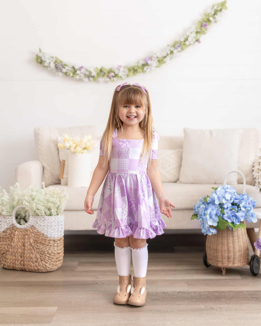 little girl surrounded by baskets of flowers in purple floral patchwork print twirl dress pared with white lace top knee high socks and lavender lace pigtail bows