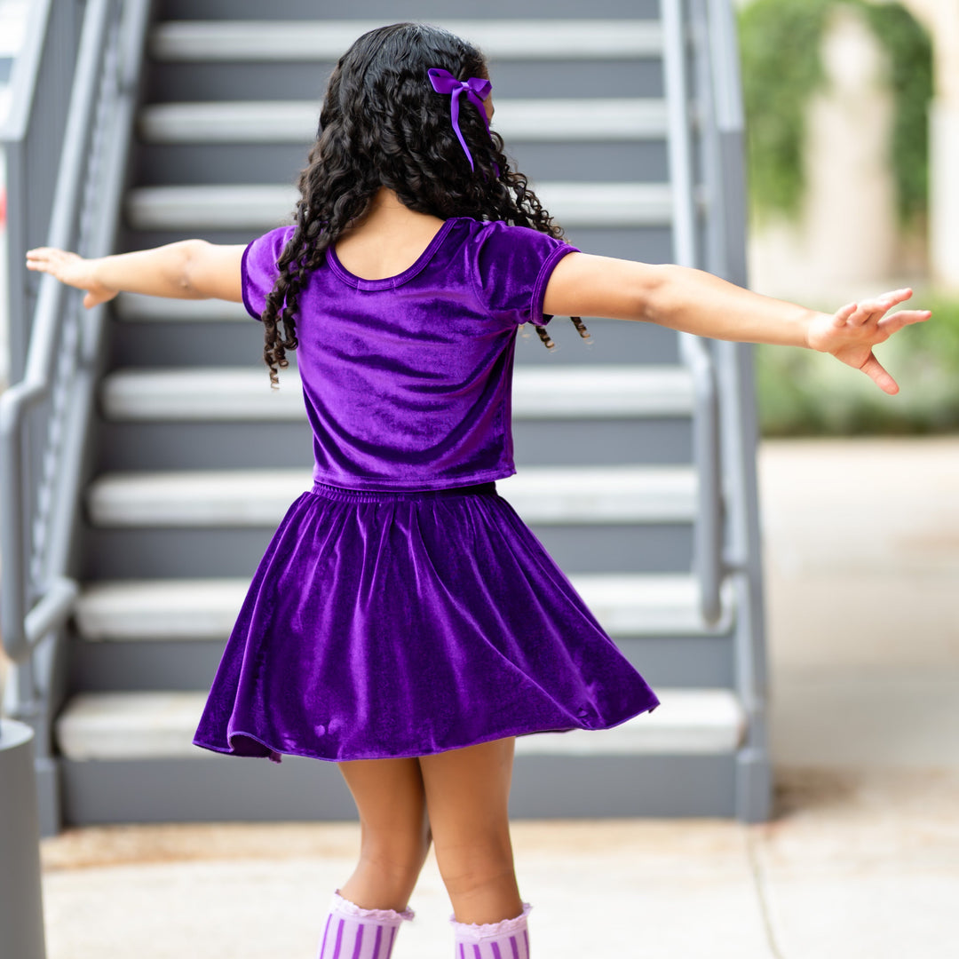 girl twirling in head to toe purple outfit including velvet top and skort, satin bow and striped lace top knee highs
