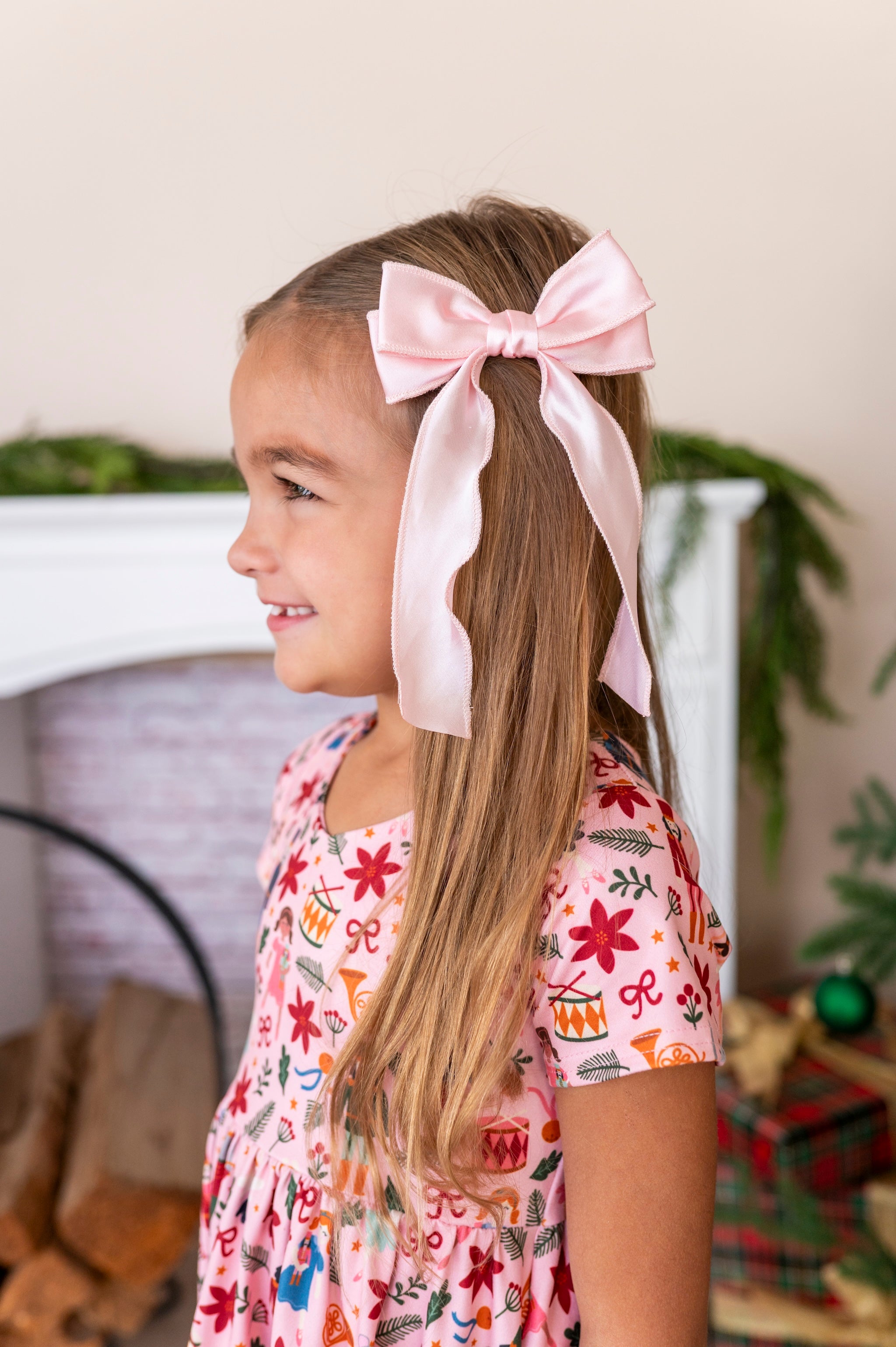 A young girl with long light brown hair wearing a large light pink satin bow in her ponytail, paired with a pink nutcracker patterned dress, standing in a cozy indoor setting.